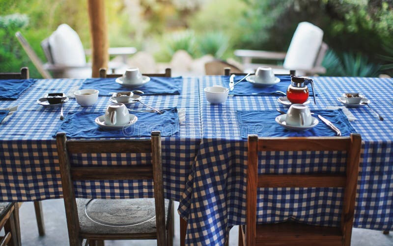 Table with empty plates and coffee cups, blue chequered tablecloth, ready for morning breakfast, blurred green foliage in background - eating at tropical holiday resort. Table with empty plates and coffee cups, blue chequered tablecloth, ready for morning breakfast, blurred green foliage in background - eating at tropical holiday resort.