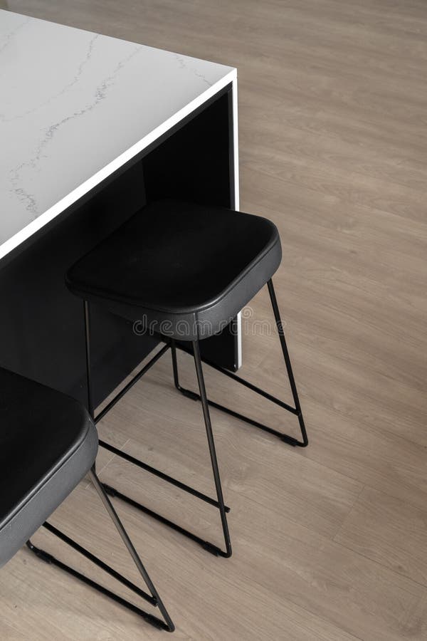 Modern styled black leather bar stools with thin metal legs underneath white kitchen counter with marble texture on linoleum floor with wooden print, vertical shot. Modern styled black leather bar stools with thin metal legs underneath white kitchen counter with marble texture on linoleum floor with wooden print, vertical shot