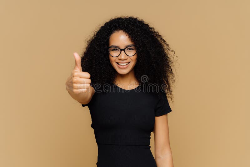 Waist up shot of satisfied supportive woman shows thumb up, cheers best friend, encourages for excellent efforts, wears glasses and black t shirt, stands against beige background. Body language. Waist up shot of satisfied supportive woman shows thumb up, cheers best friend, encourages for excellent efforts, wears glasses and black t shirt, stands against beige background. Body language