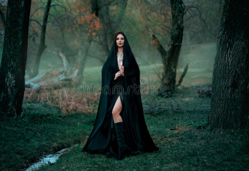Mysterious fantasy gothic woman dark witch obsessed by evil. Girl demon vampire in black dress cape hood. walk in dark dense deep forest background, trees. Medieval queen in silk cloak, scarf posing. Mysterious fantasy gothic woman dark witch obsessed by evil. Girl demon vampire in black dress cape hood. walk in dark dense deep forest background, trees. Medieval queen in silk cloak, scarf posing.