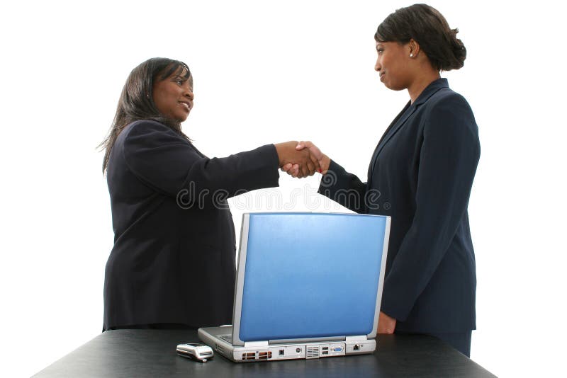 Two beautiful African American business women shaking hands. Standing behind a table with laptop and cellphone. Over white background. Women in mid and late 20's. Two beautiful African American business women shaking hands. Standing behind a table with laptop and cellphone. Over white background. Women in mid and late 20's.