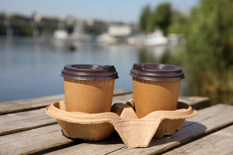 Takeaway paper coffee cups with plastic lids in cardboard holder on wooden pier near river royalty free stock photography