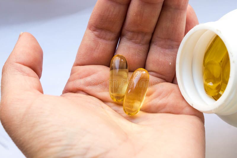 A white can of fish oil in jelly-like transparent yellow capsules and one dose two capsules of capsules in a woman`s palm close-up on a white background. A white can of fish oil in jelly-like transparent yellow capsules and one dose two capsules of capsules in a woman`s palm close-up on a white background