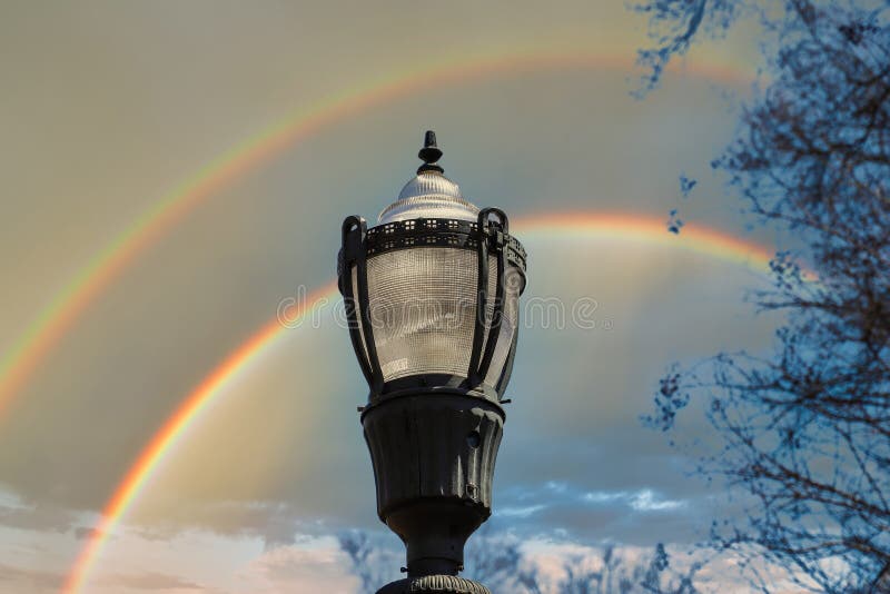 A tall black lamp post surrounded by bare winter trees with clear blue sky and a rainbow in the sky in the Marietta Square