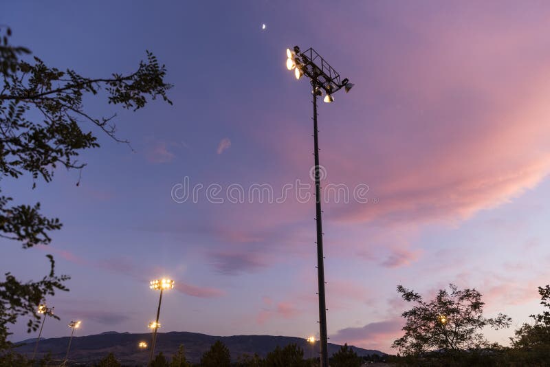 Tall Stadium Light Pole Shines Over a Baseball Field at Sunset with a Moon in the Sky Stock