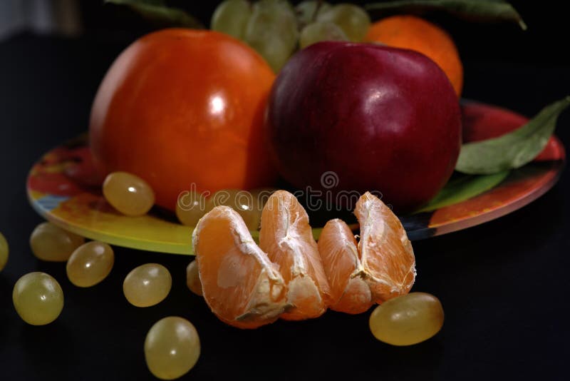 Tangerine φέτες και πράσινα σταφύλια μπροστά από ένα πιάτο με τα φρούτα σε ένα μαύρο υπόβαθρο, κινηματογράφηση σε πρώτο πλάνο στοκ φωτογραφίες με δικαίωμα ελεύθερης χρήσης Tangerine φέτες και πράσινα σταφύλια μπροστά από ένα πιάτο με τα φρούτα σε ένα μαύρο υπόβαθρο, κινηματογράφηση σε πρώτο πλάνο στοκ φωτογραφίες με δικαίωμα ελεύθερης χρήσης
