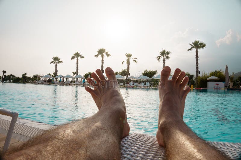 Tanned hairy men`s legs against the background of a large pool with palm trees, the concept of recreation, relaxation, tourism.