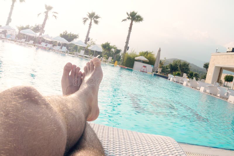 Tanned hairy men`s legs against the background of a large pool with palm trees, the concept of recreation, relaxation, tourism.