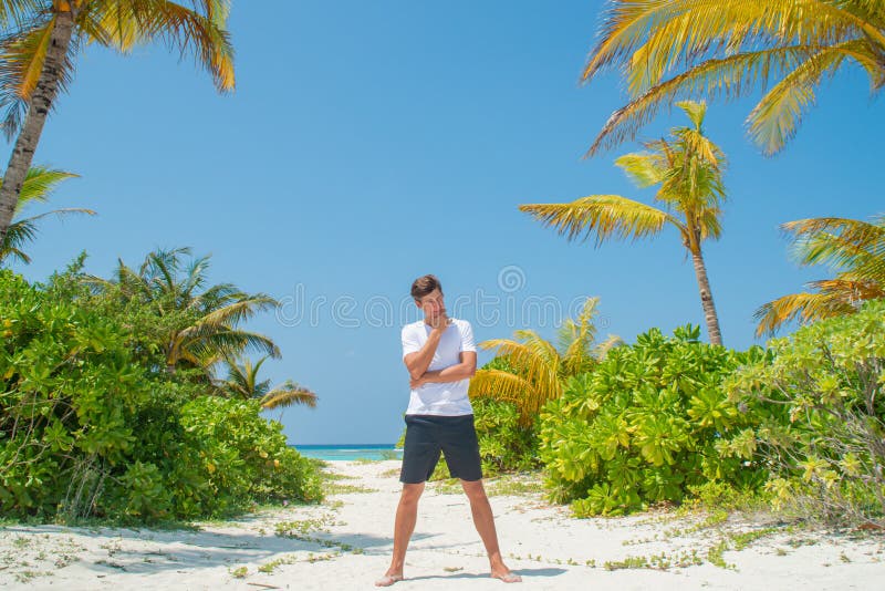 Tanned handsome young man wearing white t-shirt and black shorts standing at tropical sandy beach at island luxury resort. In Maldives royalty free stock image Tanned handsome young man wearing white t-shirt and black shorts standing at tropical sandy beach at island luxury resort. In Maldives royalty free stock image