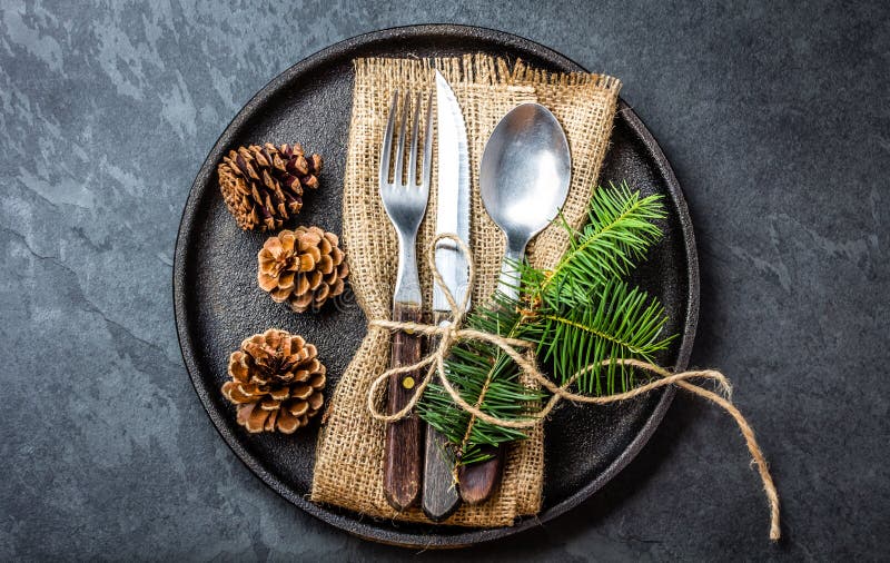 Vintage rustic cutlery set with country style napkin and Christmas decoration - fir tree brunch, cones on black iron plate on black slate background. Copy space, top view. Christmas menu background. Vintage rustic cutlery set with country style napkin and Christmas decoration - fir tree brunch, cones on black iron plate on black slate background. Copy space, top view. Christmas menu background