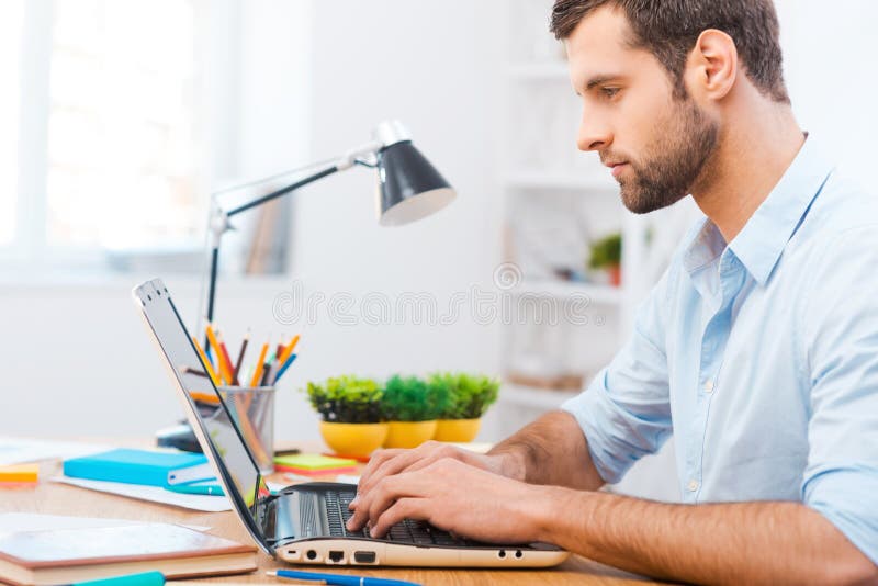 Handsome young man in shirt working on laptop while sitting at his working place. Handsome young man in shirt working on laptop while sitting at his working place