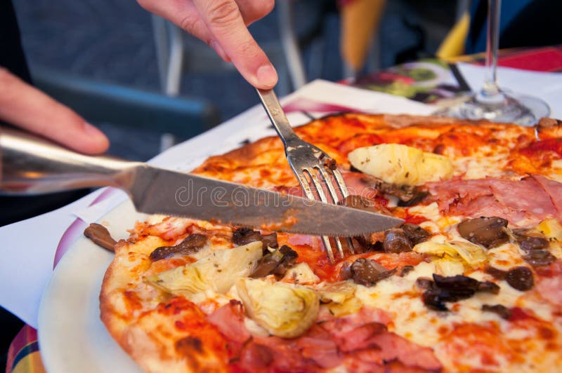 Cutting a delicious Italian pizza with cheese, mushrooms, artichokes and tomatoes with help of knife and fork. Focus on fork. Cutting a delicious Italian pizza with cheese, mushrooms, artichokes and tomatoes with help of knife and fork. Focus on fork