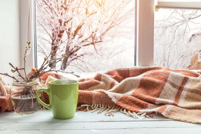 Cup of coffee, books, branch of cherry tree, wool blanket on windowsill. In the background snow tree pattern on window. Cozy home concept. Cup of coffee, books, branch of cherry tree, wool blanket on windowsill. In the background snow tree pattern on window. Cozy home concept.