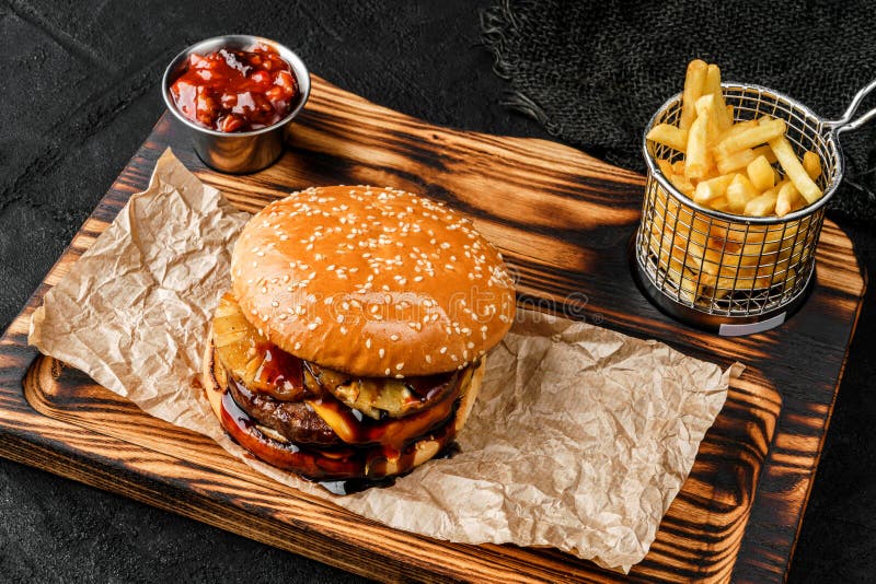 Tasty burger with slices of pineapple, meat cutlet, slice of cheese and sauce on wooden board with french fries potatoes on black