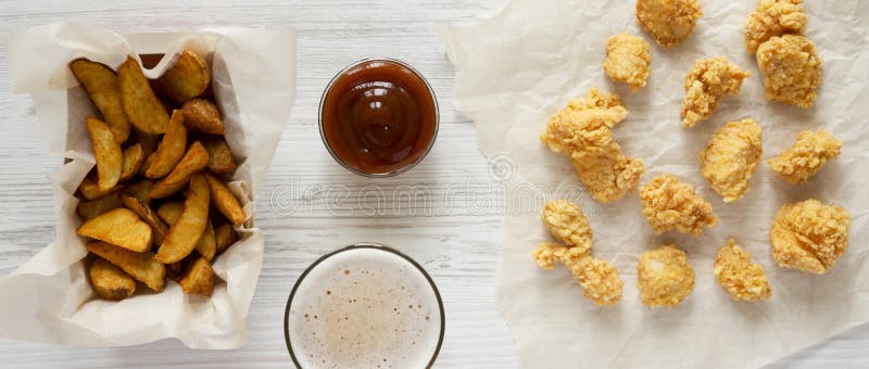 Tasty fastfood: fried potato wedges in paper box, chicken bites, barbecue sauce and beer on a white wooden table, top view. Flat