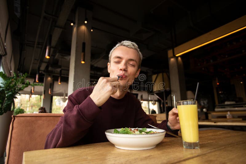 Tasty meal in a restaurant. Very emotional blond man in red pullover enjoying caesar salad and orange fresh juice