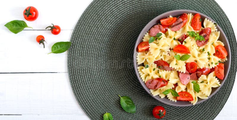 Tasty pasta farfalle with grilled sausages, fresh cherry tomatoes and basil on a plate on a white background. Top view, flat lay.