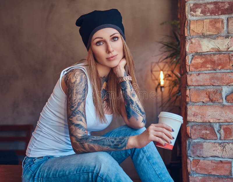 A Tattoed Blonde in a T-shirt and a Hat with Cup of Coffee in a Room ...