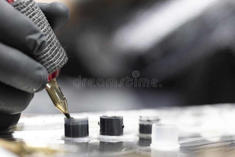 Detail of the tattooist's hand with black glove holding the tattoo machine while he loads ink into the needle while performing a tattoo. Image in horizontal and colour. Detail of the tattooist's hand with black glove holding the tattoo machine while he loads ink into the needle while performing a tattoo. Image in horizontal and colour