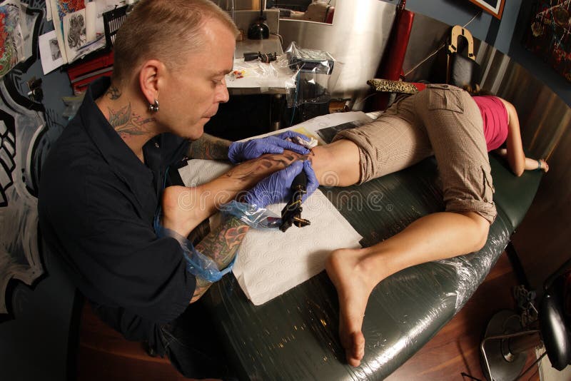 A tattoo artist applying his craft onto the lower leg of a pain-inflicted female. (Property release supplied includes tattooists' wall mural and drawings posted on the walls behind him.)