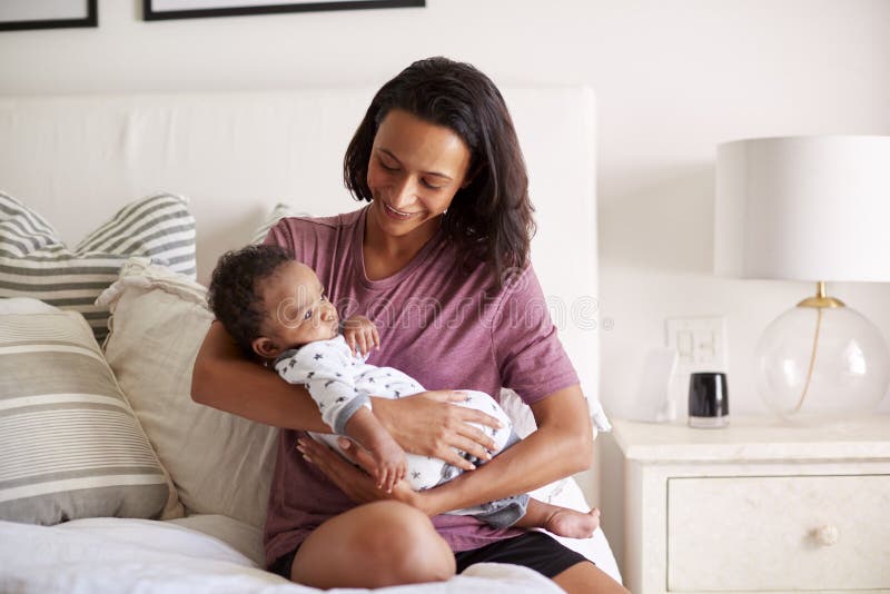 Tausendjährige Afroamerikanermutter, die auf ihrem Bett hält ihren dreimonatigen alten Babysohn in ihren Armen sitzt lizenzfreies stockbild