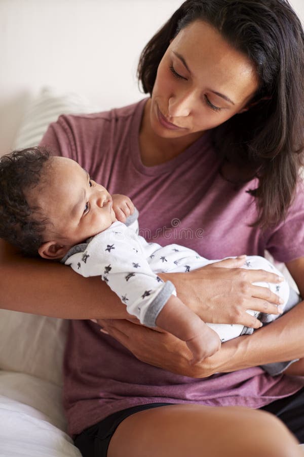 Tausendjährige Afroamerikanermutter, die auf ihrem Bett hält ihren dreimonatigen alten Babysohn in ihren Armen, vertikal sitzt stockfoto