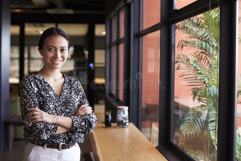 Tausendjährige asiatische Geschäftsfrau, die das Fenster in einem Büro oben lächelt zur Kamera, Taille bereitsteht lizenzfreie stockfotos Tausendjährige asiatische Geschäftsfrau, die das Fenster in einem Büro oben lächelt zur Kamera, Taille bereitsteht lizenzfreie stockfotos