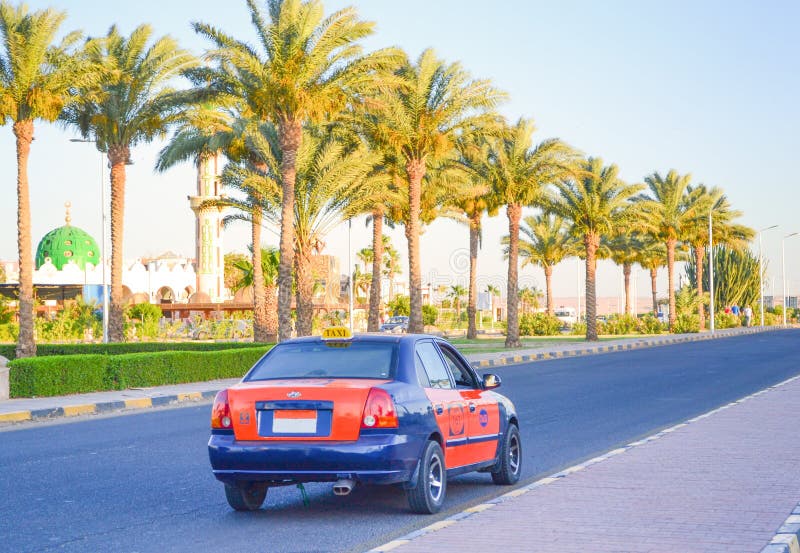 Egyptian taxi blue-orange colour goes on the road at the resort area of Hurghada. A sidewalk, a mosque and palm trees along the road in the background. Egyptian taxi blue-orange colour goes on the road at the resort area of Hurghada. A sidewalk, a mosque and palm trees along the road in the background.