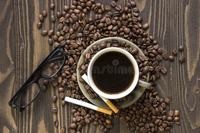 Cup of coffee with beans, two cigarettes and glasses standing on a wooden table. Top view. Cup of coffee with beans, two cigarettes and glasses standing on a wooden table. Top view