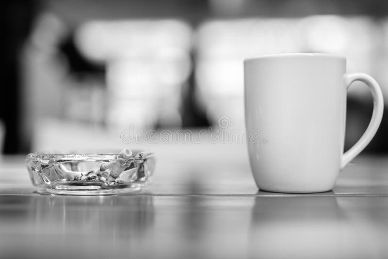 Coffee cup and ashtray with cigarettes on wooden table inside the restaurant in black and white. Coffee cup and ashtray with cigarettes on wooden table inside the restaurant in black and white