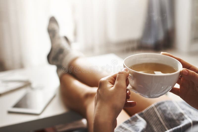 Taza de té y de frialdad Mujer que miente en el sofá, llevando a cabo las piernas en la mesa de centro, el café caliente de consu foto de archivo