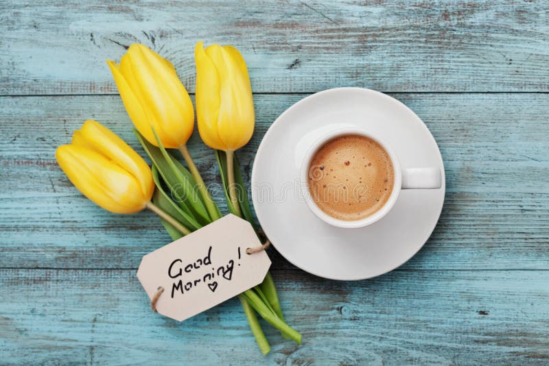 Tazza da caffè con il buongiorno giallo dei fiori e delle note del tulipano sulla tavola rustica blu da sopra