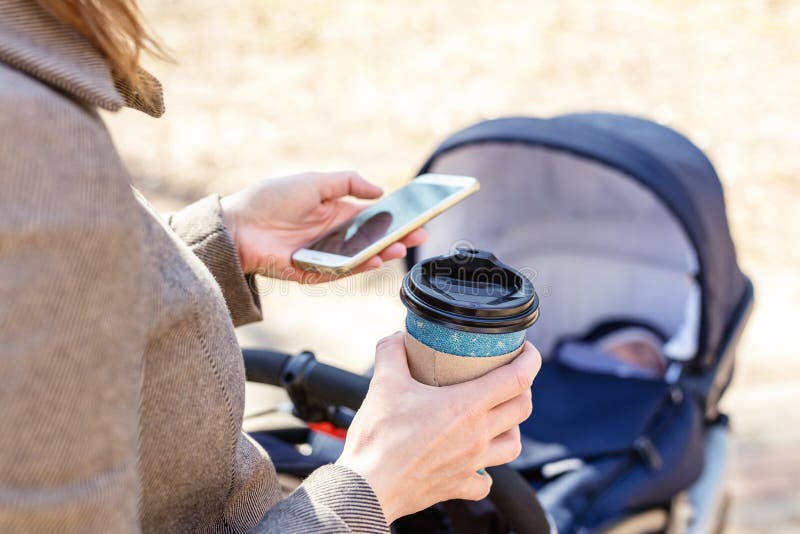 Tazza di caffè della tenuta della donna da andare per mezzo dello smartphone durante la passeggiata con carrozzina Giovane donna immagini stock Tazza di caffè della tenuta della donna da andare per mezzo dello smartphone durante la passeggiata con carrozzina Giovane donna immagini stock
