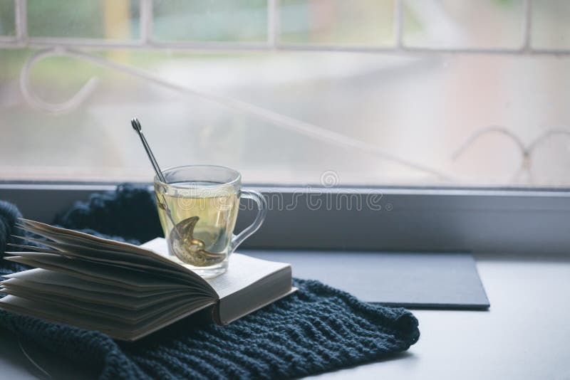 Tazza tricottata del maglione del libro aperto caldo del tè sulla finestra Giorno accogliente di autunno di inverno Atmosfera tra immagini stock libere da diritti Tazza tricottata del maglione del libro aperto caldo del tè sulla finestra Giorno accogliente di autunno di inverno Atmosfera tra immagini stock libere da diritti