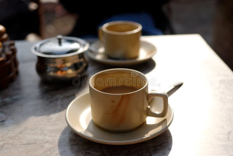 A closeup of two beige teacups, filled with Indian tea (called chae). They are placed on a table, with a small jar of sugar nearby. A closeup of two beige teacups, filled with Indian tea (called chae). They are placed on a table, with a small jar of sugar nearby.