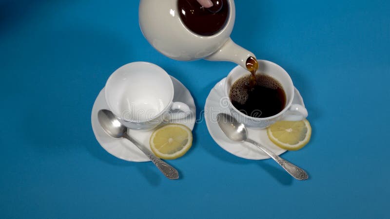 Tea is poured from a white teapot into white porcelain cups with lemon slices on saucers. Tea still life on a blue