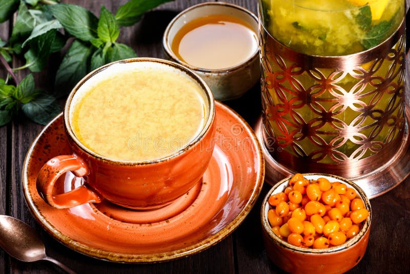 Tea with sea Buckthorn in cup with saucer, served with vintage teapot, fresh berries, honey, mint on dark wooden background