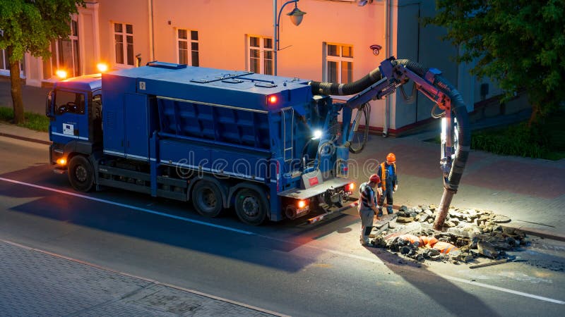 A team of road builders perform road repairs at night with the help of a large truck vacuum cleaner. Professional repairmen change