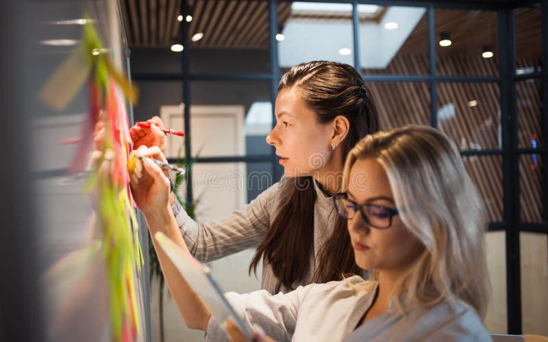 Teamwork, project management, agile methodology. Two young business women in the office are planning product development and support. Colleagues glue sticky stickers on the Board. Software methodology stock images, royalty-free photos and pictures