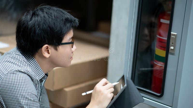 Young Asian male technician checking red fire extinguisher in fire hose cabinet FHC by using digital tablet and pen. Building service and maintenance concepts. Young Asian male technician checking red fire extinguisher in fire hose cabinet FHC by using digital tablet and pen. Building service and maintenance concepts