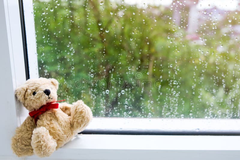 Teddy Bear con el arco rojo de la cinta se sienta tristemente por la ventana y el exterior, él el ` s que llueve y frío fotos de archivo Teddy Bear con el arco rojo de la cinta se sienta tristemente por la ventana y el exterior, él el ` s que llueve y frío fotos de archivo