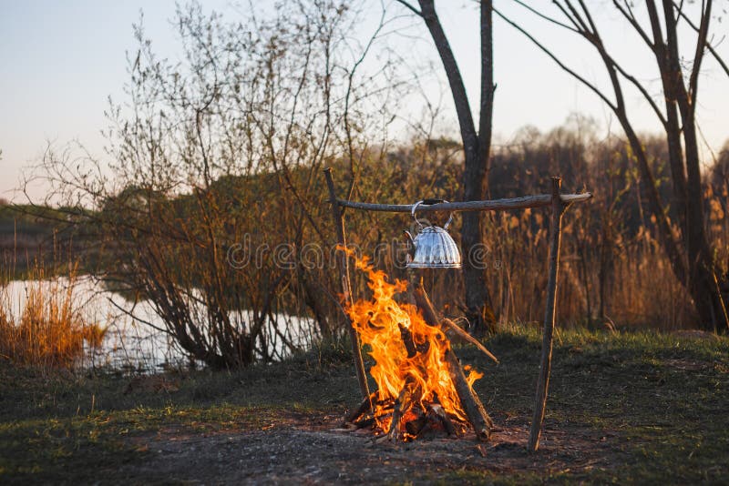 Teekanne mit Tee h?ngt ?ber dem Feuer auf dem Riverbanksonnenuntergang lizenzfreie stockbilder Teekanne mit Tee h?ngt ?ber dem Feuer auf dem Riverbanksonnenuntergang lizenzfreie stockbilder