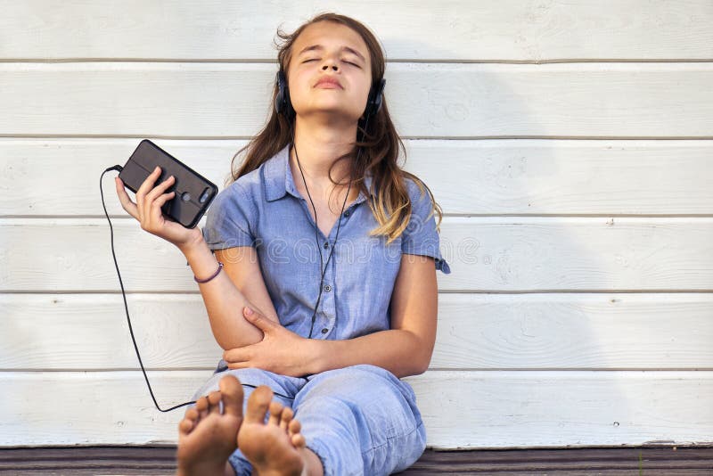 Teen barefoot girl wearing headphones enjoying music from a smartphone and singing outdoors in summer evening