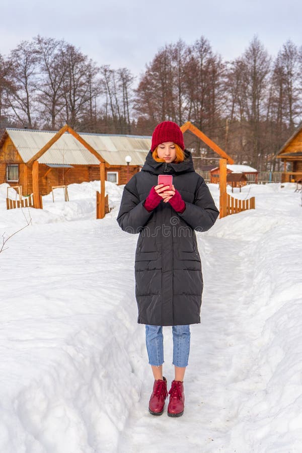 Teen cute girl in black down jacket, blue cropped jeans, burgundy hat, boots and fingerless gloves standing outdoor against winter