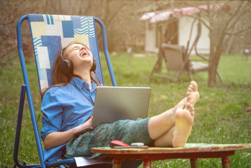 Teen girl barefoot laughing during online conference with her school friends sitting in a deckchair outdoors in springtime