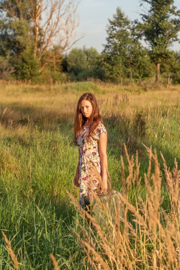 A young girl in a retro vintage dress with flowing hair is standing in a tall grass in a sunlight afterglow with a suitcase