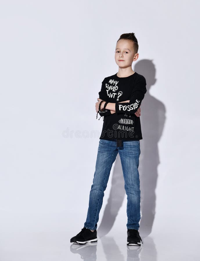 Teenage boy in black jumper and sneakers, blue jeans, bracelet. He standing with folded hands, posing isolated on white background