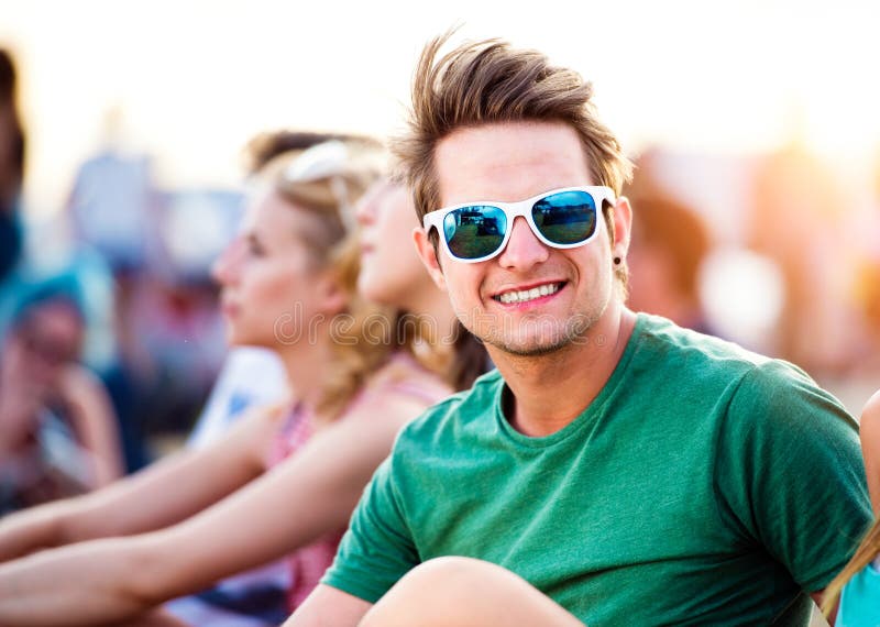 Hipster teenage boy in green t-shirt and sunglasses with his friends at summer music festival, sitting on the ground. Hipster teenage boy in green t-shirt and sunglasses with his friends at summer music festival, sitting on the ground