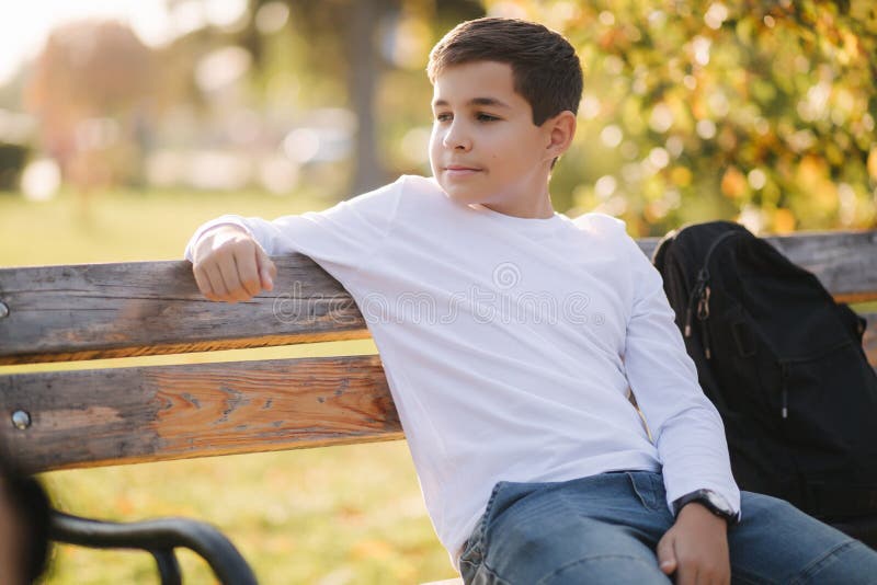 Teenage boy in white sweater sitting on the bench. Handsome school boy with backpack in autumn park stock image Teenage boy in white sweater sitting on the bench. Handsome school boy with backpack in autumn park stock image