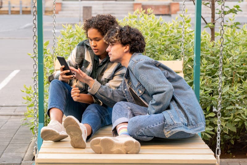 Teenage couple sitting on swings and scrolling in smartphone